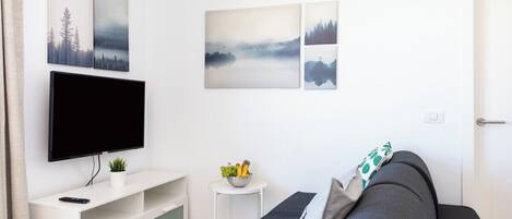 A modern living room with a flat-screen TV on a white stand, a dark grey sofa, and a small white side table with a bowl of fruit.