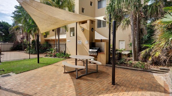 An outdoor patio area with a barbecue grill, a table and benches, surrounded by palm trees and a brick floor.
