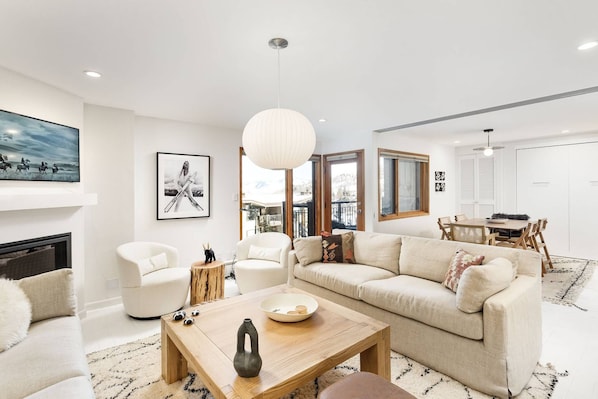 A modern living room with a sofa, coffee table, and a fireplace. There is a dining area with a table and chairs.