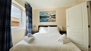 A neatly made bed with white linens, a painting of a mountain landscape on the wall, and a window with curtains. - Broad Gauge Apartments (Wolverhampton)