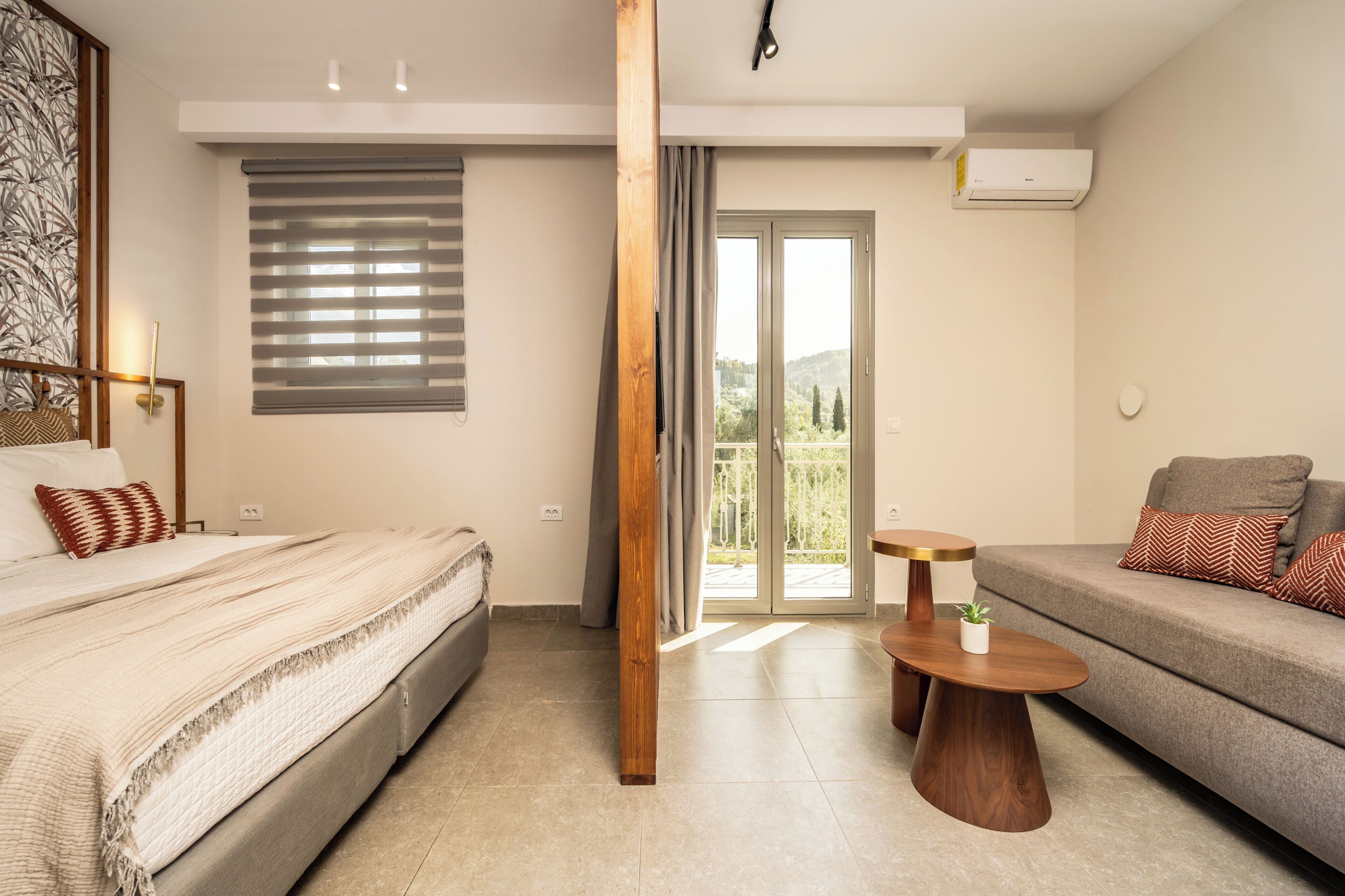 A modern hotel room with a bed, sofa, wooden floor, and a view through a glass door.