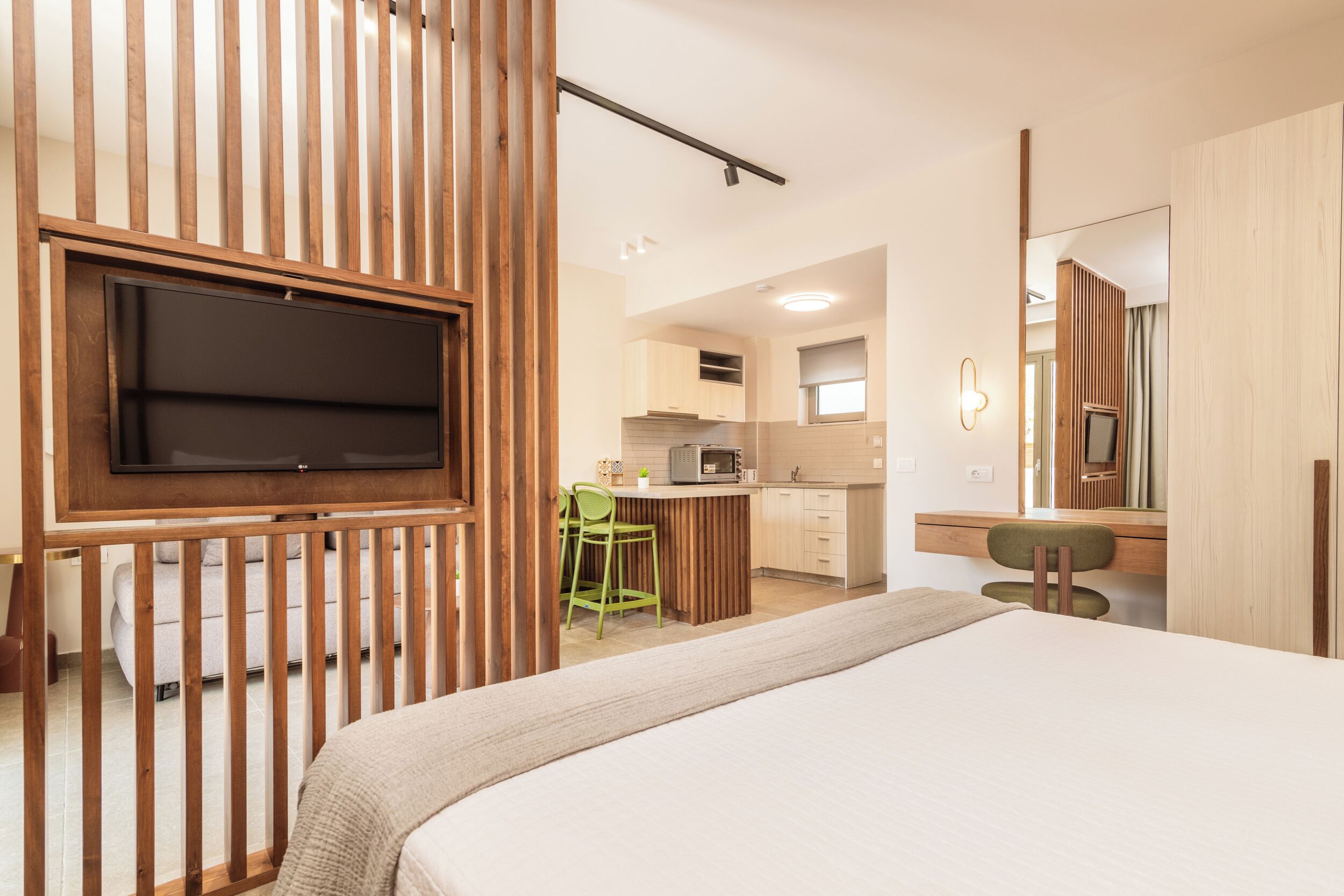 A bedroom with a bed, a TV mounted on a wooden panel, a kitchenette, and a bathroom with a mirror.
