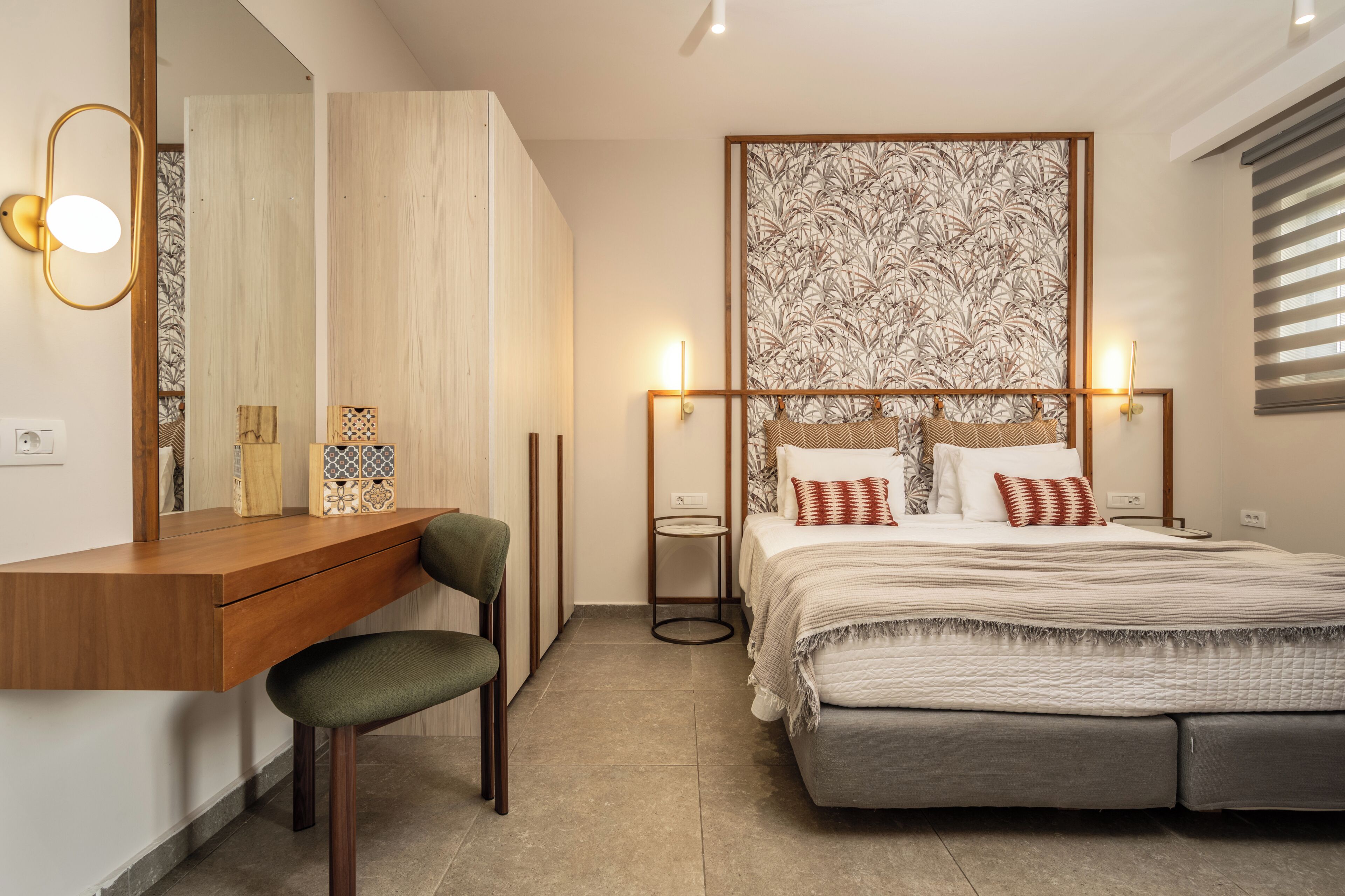 A bedroom with a wooden desk, a chair, a bed with a patterned headboard, bedside tables, and a large mirror.