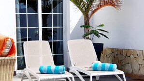 Two white lounge chairs with blue cushions, a wicker basket, and a potted plant on a patio with a stone wall and a glass door. - Spacious three bedroom holiday home with pool (La Caleta)
