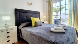A neatly made bed with a grey quilt, a dark headboard, and two pillows. There is a white bedside table with a lamp and a drawer. A window with curtains is visible, and a chair is seen outside. - Spacious three bedroom holiday home with pool (La Caleta)