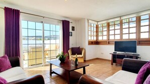 A living room with a sofa, armchair, and a table with a bottle and glasses. There is a TV on a stand and a window with curtains. - Spacious three bedroom holiday home with pool (La Caleta)