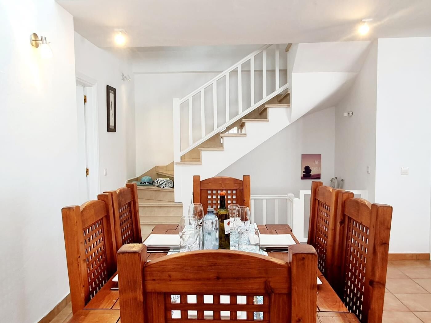 A dining area with a wooden table set for four, a staircase in the background, and a 'Live your best life' poster on the wall.