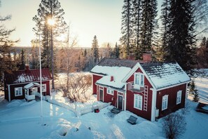Una casa de madera roja con techo nevado, rodeada de altos pinos y un cielo despejado.