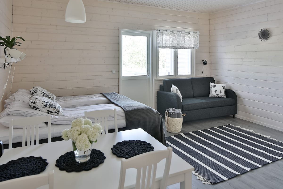 Ein Zimmer mit einem Bett, einer Couch, einem Esstisch mit Blumenvase und einem Fenster mit Vorhängen.