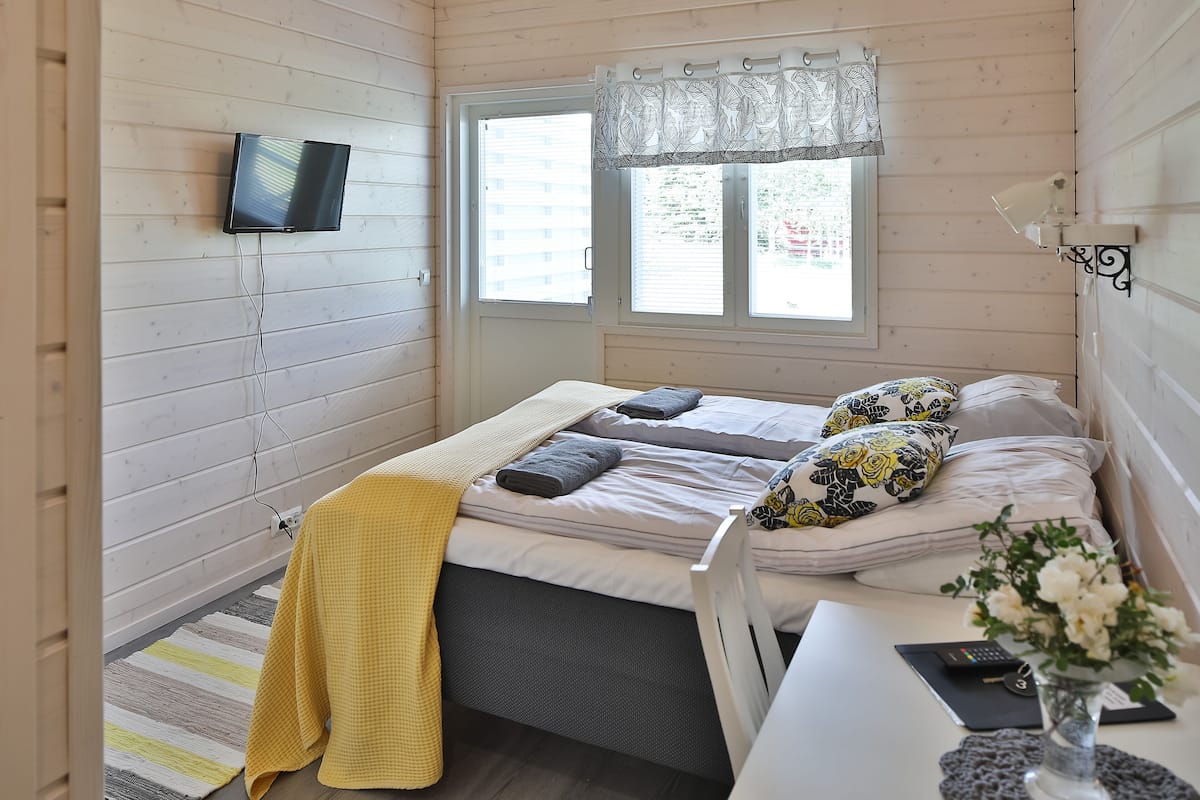 Ein Schlafzimmer mit einem Bett, einem an der Wand befestigten Fernseher, einem Fenster mit Vorhängen und einem Nachttisch mit Blumen.
