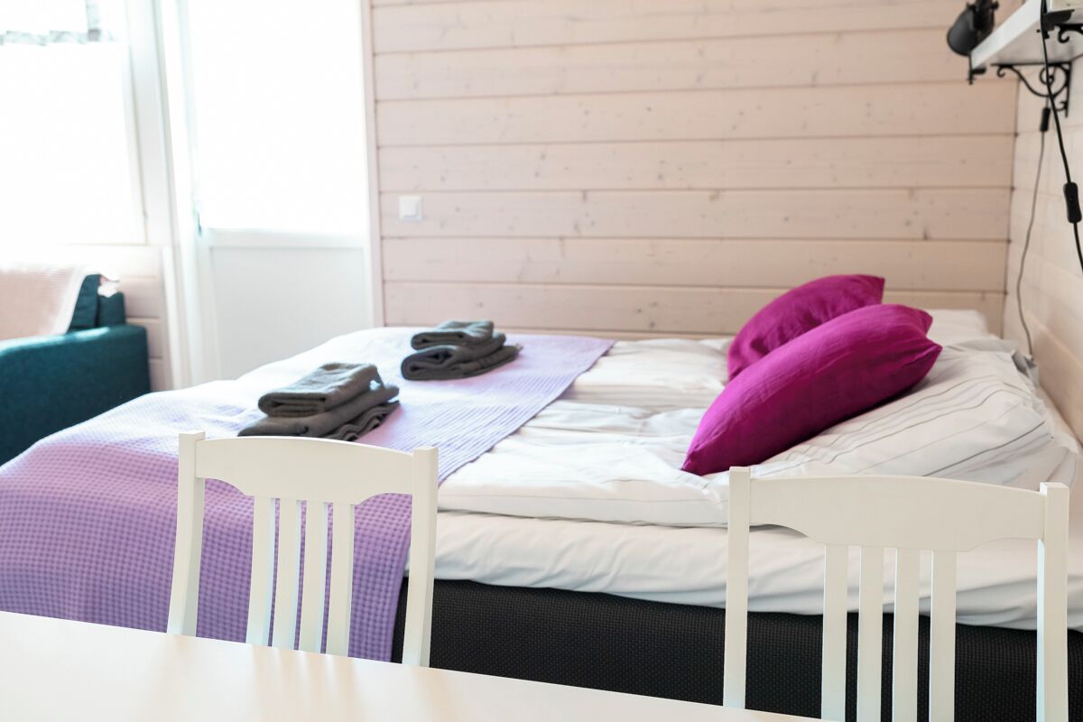 A neatly made bed with a purple towel, a pair of slippers, and a pink pillow. There is a white chair in front of the bed, and a table with a purple checkered tablecloth.