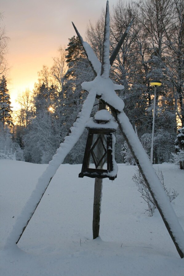 Una estructura de madera cubierta de nieve con un farol, rodeada de árboles y un atardecer al fondo.