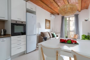Une cuisine moderne avec des armoires blanches, un four encastré et un réfrigérateur blanc. On y trouve une table à manger avec des chaises et un luminaire suspendu en osier.