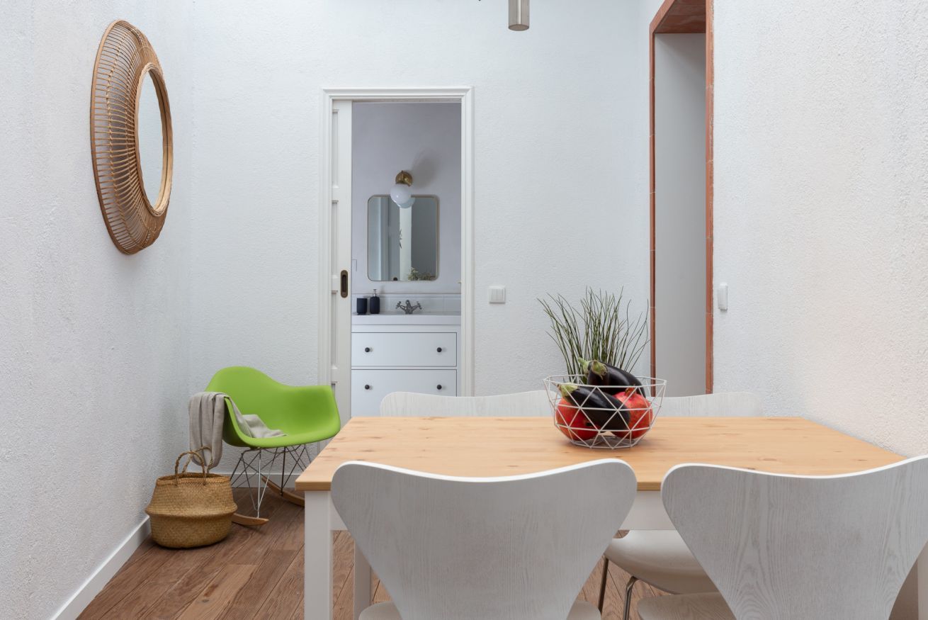 Un comedor con una mesa de madera, sillas blancas, una silla verde, una cesta de mimbre y un bol de metal con plantas.