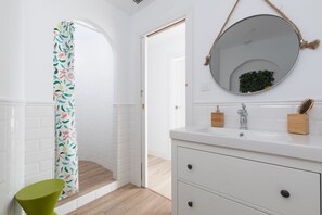 A bathroom with a white brick wall, a white vanity with a round mirror, and a shower curtain with a floral pattern. - Casa Cosi - Eixample 3 (Barcelona)