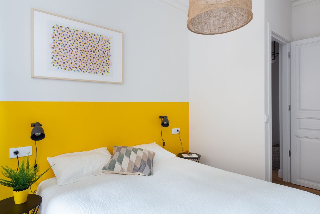 Un dormitorio con pared de acento amarilla, una cama con ropa de cama blanca, un cuadro abstracto enmarcado, una lámpara colgante y una mesita con una planta.