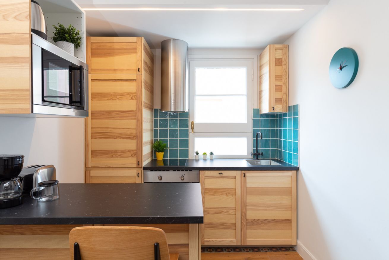Een kleine keuken met houten kastjes, een zwart aanrecht, een rvs afzuigkap en een klok aan de muur.