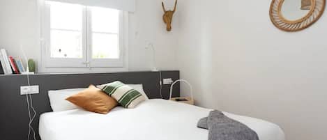 Ein Schlafzimmer mit einem Bett, einem Fenster, einem wandmontierten Spiegel und einem Wandbild.