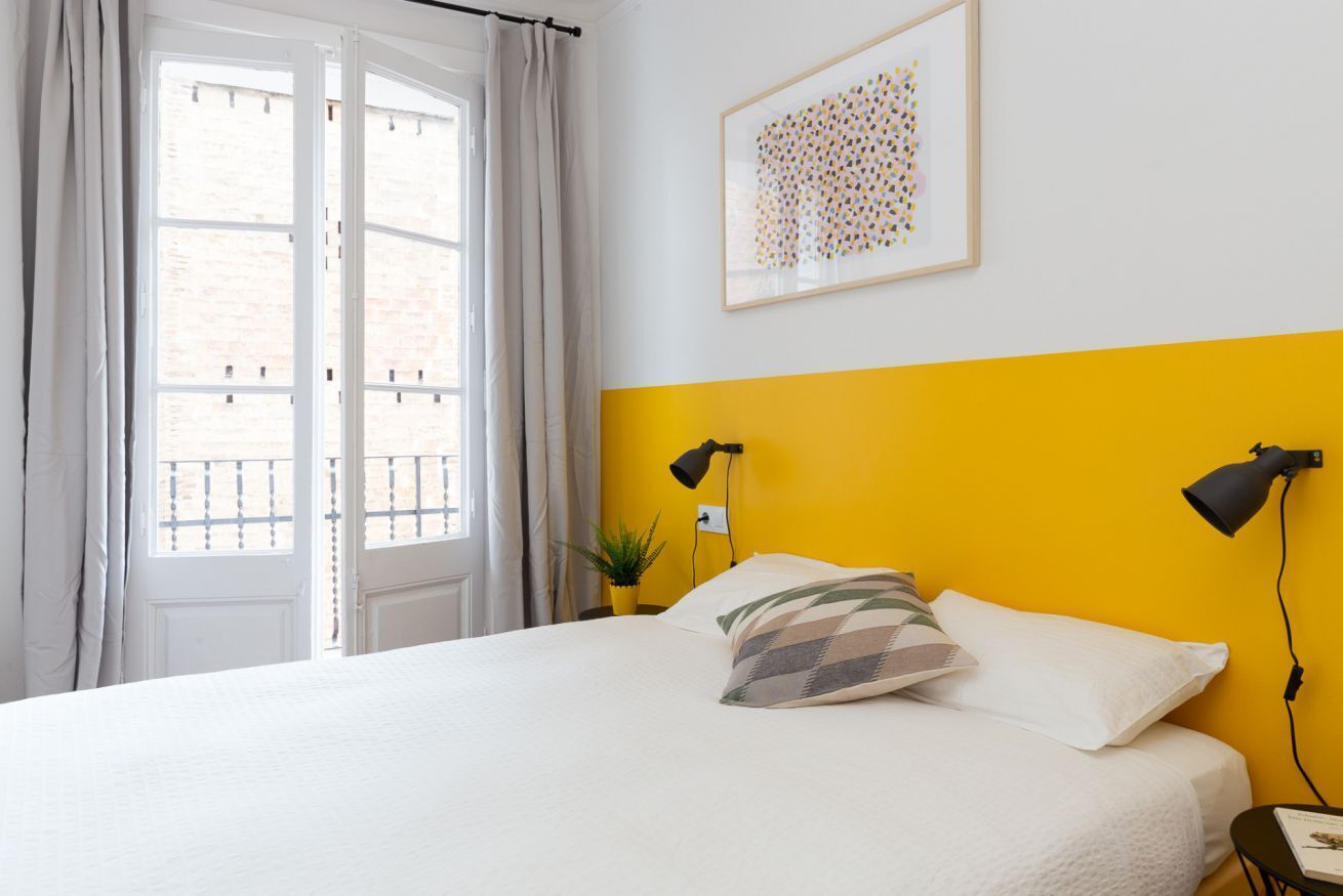 Een slaapkamer met een bed, een raam met gordijnen, een geel accentmuur, een ingelijst kunstwerk en twee bedlampen.