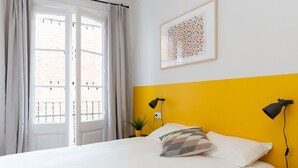 A bedroom with a bed, a window with curtains, a yellow accent wall, a framed artwork, and two bedside lamps.