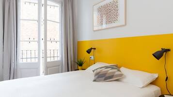 Een slaapkamer met een bed, een raam met gordijnen, een geel accentmuur, een ingelijst kunstwerk en twee bedlampen.
