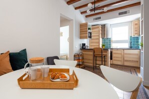 Eine moderne Küche mit Holzschränken, ein Essbereich mit weißem Tisch und Stühlen sowie ein Frühstücksbrett mit Croissants.