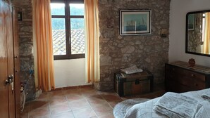 A room with a stone wall, a window with curtains, a bed with a patterned blanket, a wooden cabinet, and a small chest. - Family apartment located between the Priorat wine district and the beach in Cambrils (Duesaigües)