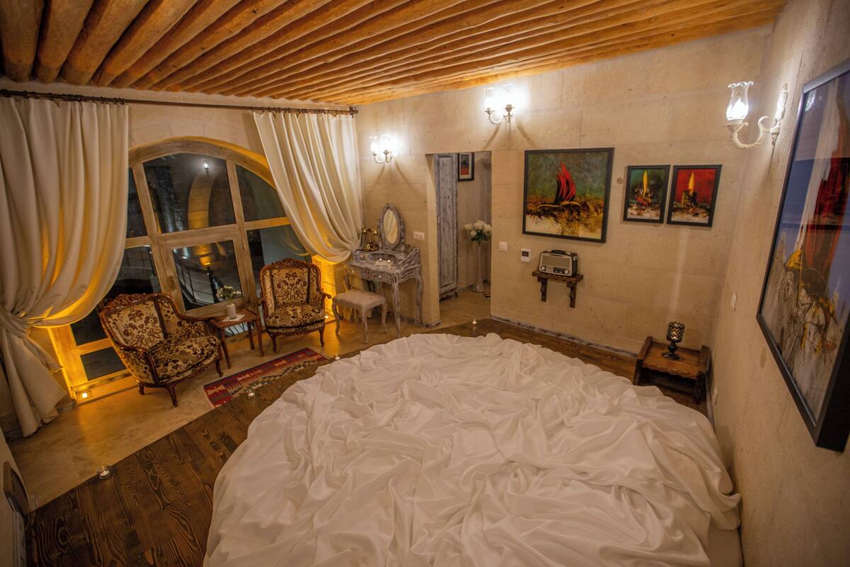 A bedroom with a large bed, a sitting area with a chair and table, a mirror, and artwork on the walls.
