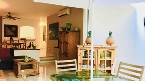 Un coin repas avec une table à plateau de verre, des chaises en bois et un parasol blanc. On y trouve également un meuble en bois orné d’objets décoratifs et un ventilateur de plafond.
