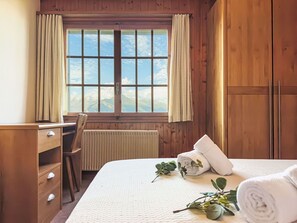 Relax in this serene bedroom with a large window offering a view of the outdoors, a comfortable bed, and a wooden desk for work.