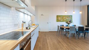 Una cocina moderna con azulejos blancos tipo metro, encimera de madera y horno empotrado. Hay una zona de comedor con mesa y sillas de madera, y un cuadro abstracto grande en la pared.
