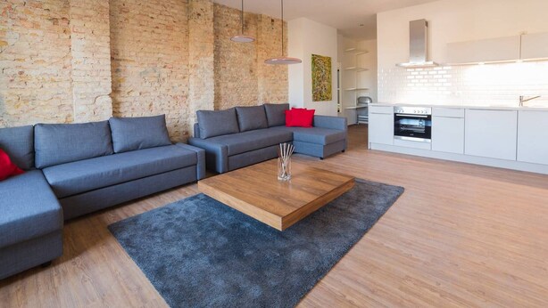 Una sala de estar moderna con un sofá modular, una mesa de centro y una zona de cocina con armarios blancos y una pared de ladrillo.