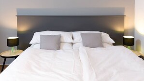 A neatly made bed with white bedding and two pillows, flanked by two bedside tables with lamps, in a modern room with a dark headboard and a light-colored wall.