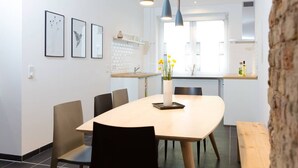 A modern dining area with a wooden table, black chairs, and a vase with yellow flowers. There are three framed pictures on the wall and a kitchen area in the background.