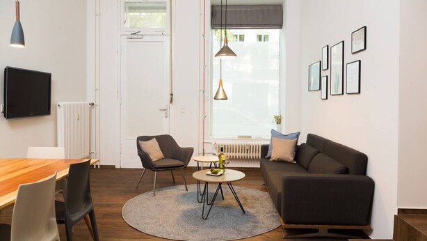 A modern living room with a wooden dining table, two chairs, a round coffee table, a sofa, and a TV mounted on the wall.
