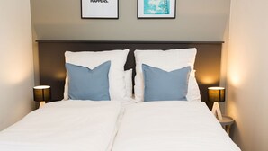 A double bed with white and blue pillows, two bedside lamps, and two framed pictures on the wall.