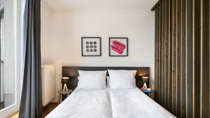 A double bed with white linens, two bedside tables with lamps, a radiator, and two framed artworks on the wall.
