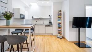 A modern kitchen with white cabinets, a wooden countertop, and a built-in oven. There is a bar area with stools and a vase of flowers. A tall stack of books is next to a flat-screen TV.
