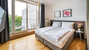 A modern bedroom with a large bed, a TV, a bedside table with a lamp, and a sliding glass door leading to a balcony.