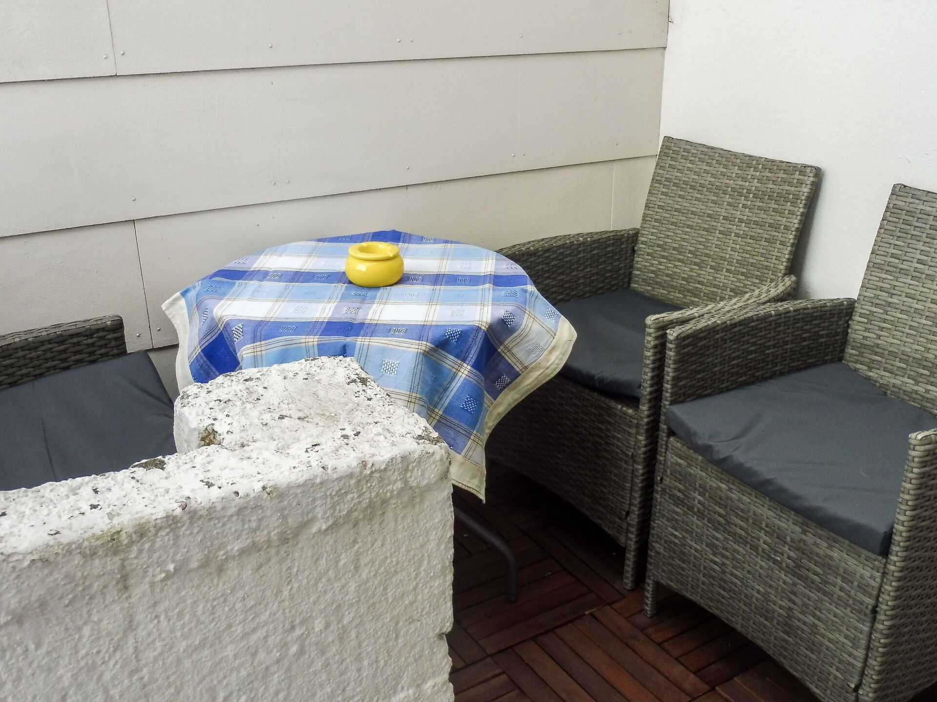 Un pequeño espacio al aire libre con una mesa cubierta con mantel a cuadros, sillas de mimbre y una jardinera de hormigón.