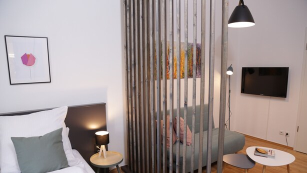 Une chambre d’hôtel moderne comprenant un lit, un canapé, une petite table, un téléviseur et un élément de séparation décoratif.