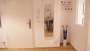 Un couloir avec une porte blanche, une petite fenêtre ornée d’un cadre, une table en bois avec des chaises, et un porte-parapluies fixé au mur.