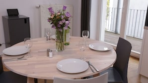 A round dining table with white plates, cutlery, and wine glasses, set against a light wooden backdrop. A vase with purple flowers is centered on the table.