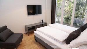 A modern bedroom with a bed, a grey sofa, a TV mounted on the wall, and a window with a view of greenery.