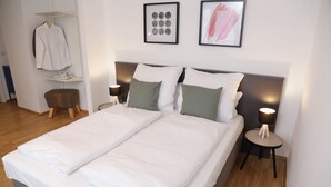 Un lit double avec du linge de lit blanc, deux tables de chevet avec des lampes, un tabouret en bois et une chemise blanche suspendue à un porte-vêtements.