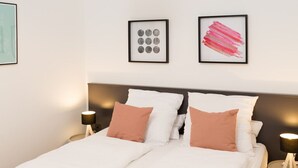 Una cama doble con almohadas blancas y melocotón, un cabecero gris y una mesita de noche con una lámpara. Hay dos obras de arte enmarcadas en la pared.