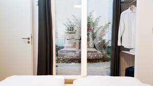 Habitación de hotel con cama, armario y vista a través de un ventanal a un área exterior con plantas y un banco.