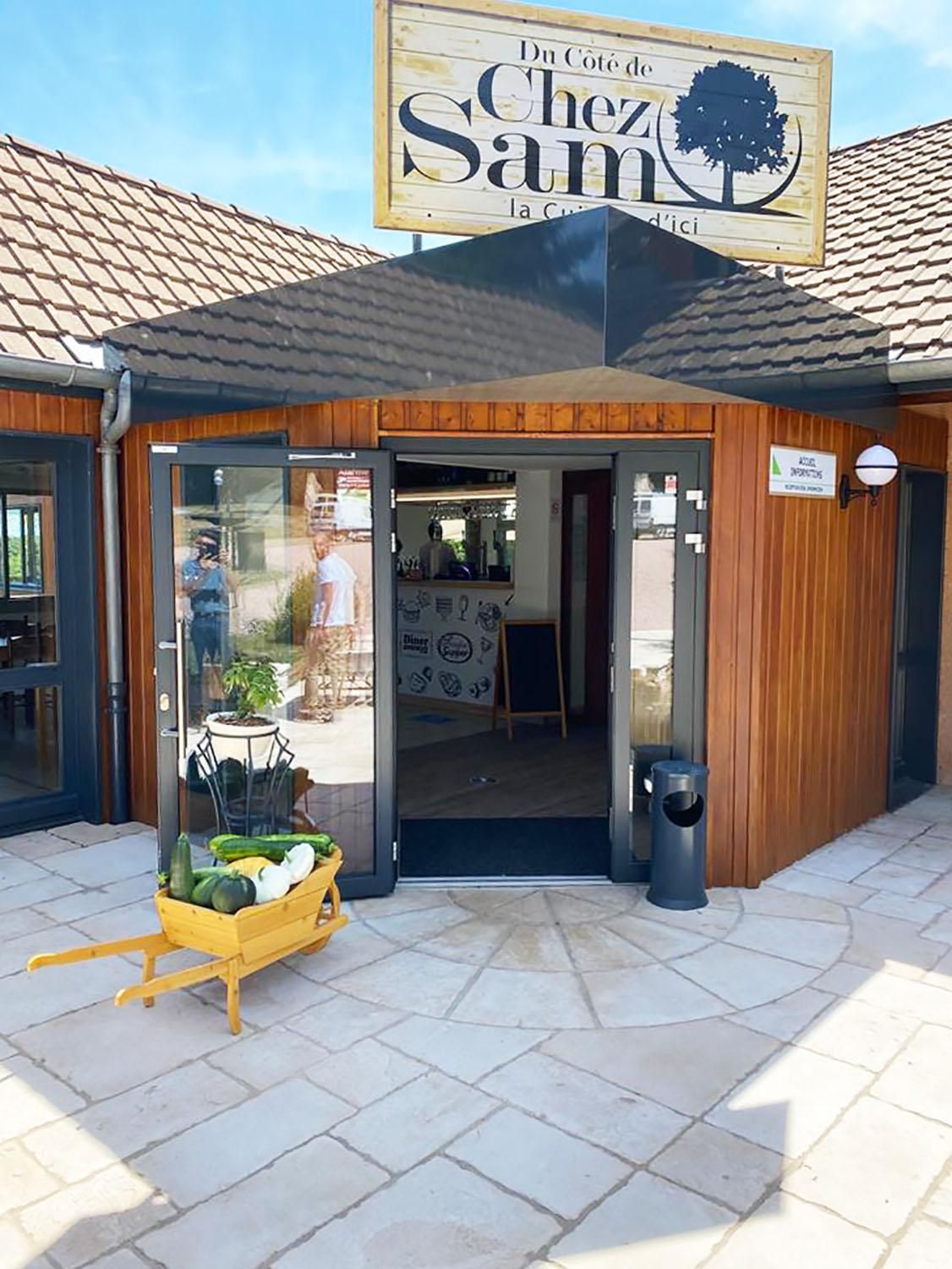Een winkelgevel met een bord waarop 'Du Côte de Chez Sam' staat en een klein terras met een houten tafel en een mand vol groenten.