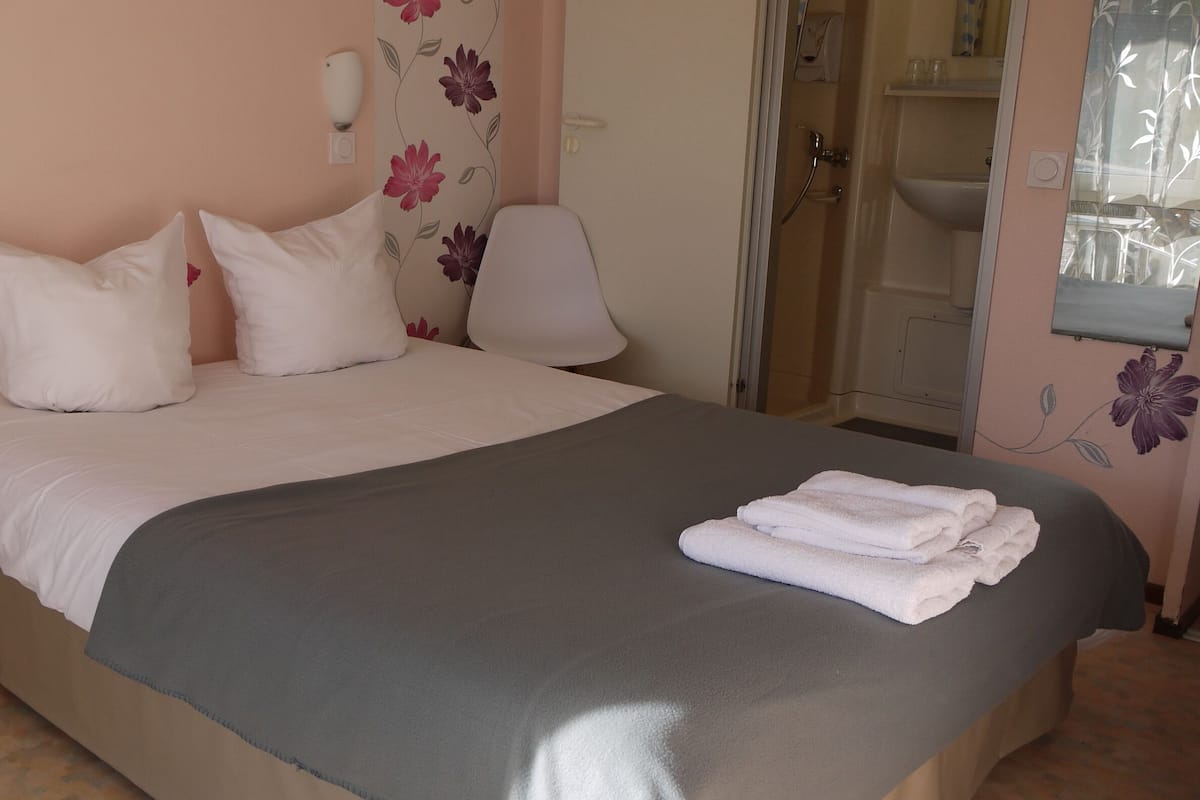 Een net opgemaakt bed met witte lakens en een grijze deken, een nachtkastje, en een badkamer met een douche en een spiegel.