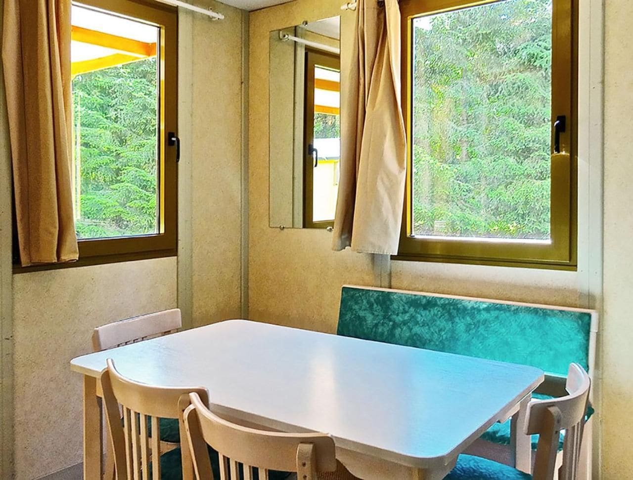 Une pièce avec une table et des chaises, une fenêtre avec des rideaux et une vue sur la verdure à l’extérieur.